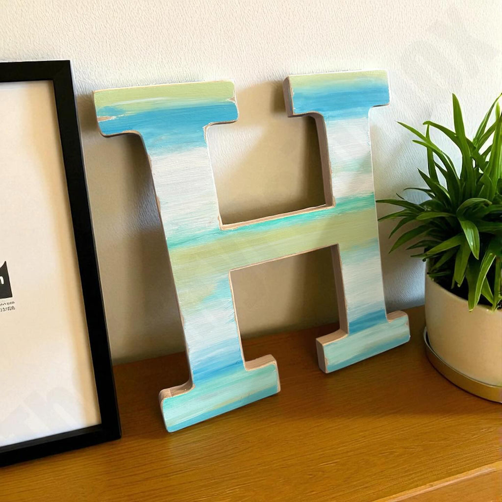 Decorative letter 'H' with a watercolor design on a wooden surface next to a plant and framed picture.